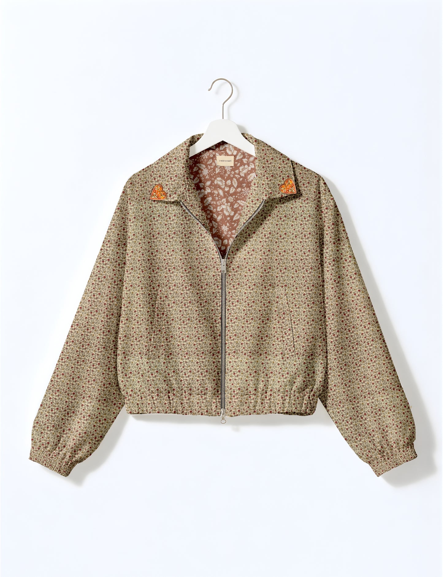 Beige floral bomber jacket with front zipper and orange heart patches on collar, hanging on white hanger
