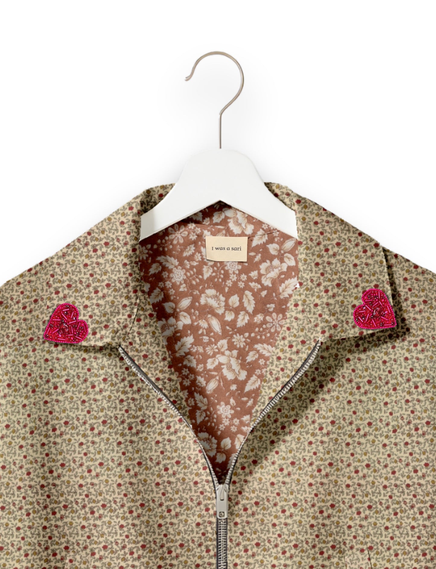 Beige floral bomber jacket with pink beaded heart patches on collar, silver zipper, and floral inner lining on white hanger