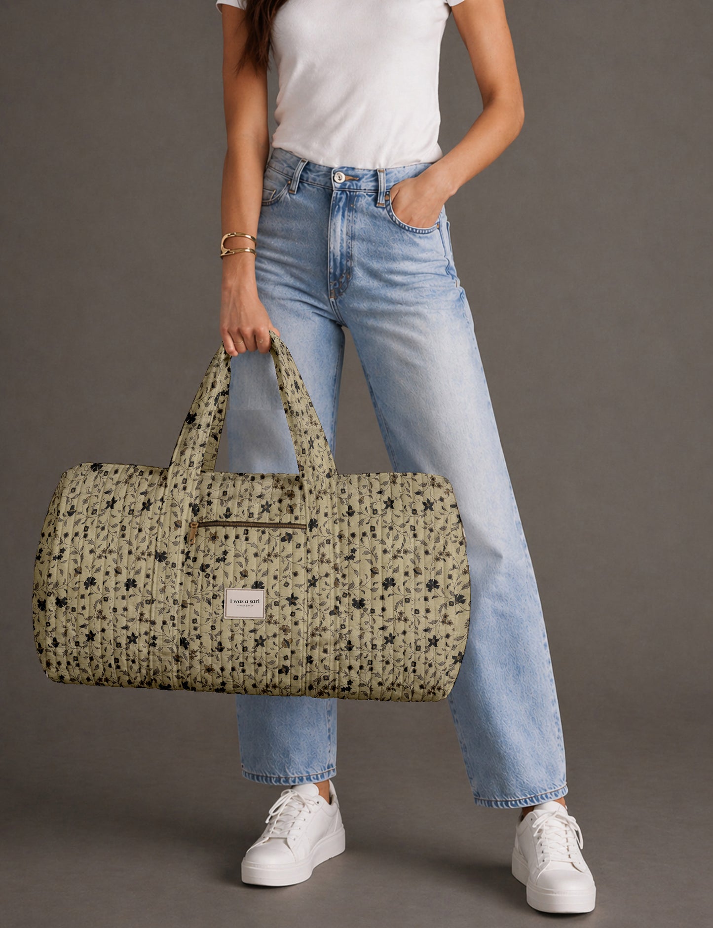 Woman holding a brown paisley quilted barrel bag with front zipper pocket, wearing light blue jeans and white sneakers