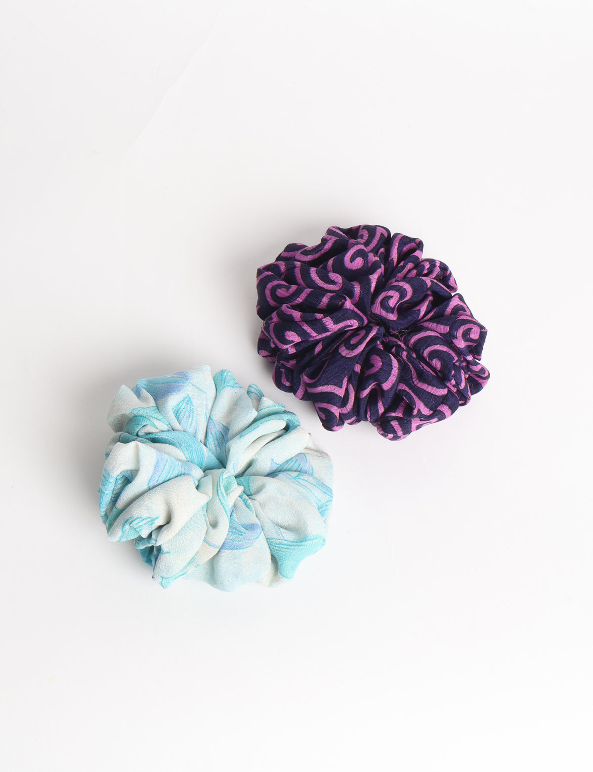 Set of two fabric scrunchies, one turquoise with beige abstract spots, one red with white linear pattern, flat lay on white
