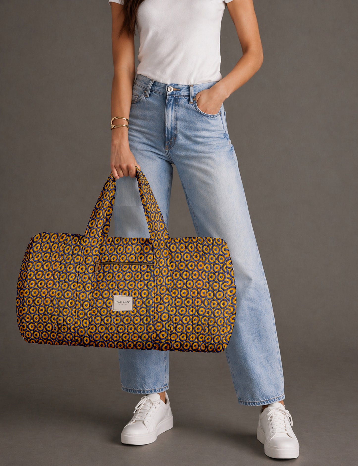 Woman holding yellow geometric patterned quilted barrel bag with front zipper pocket and label, wearing white top and light