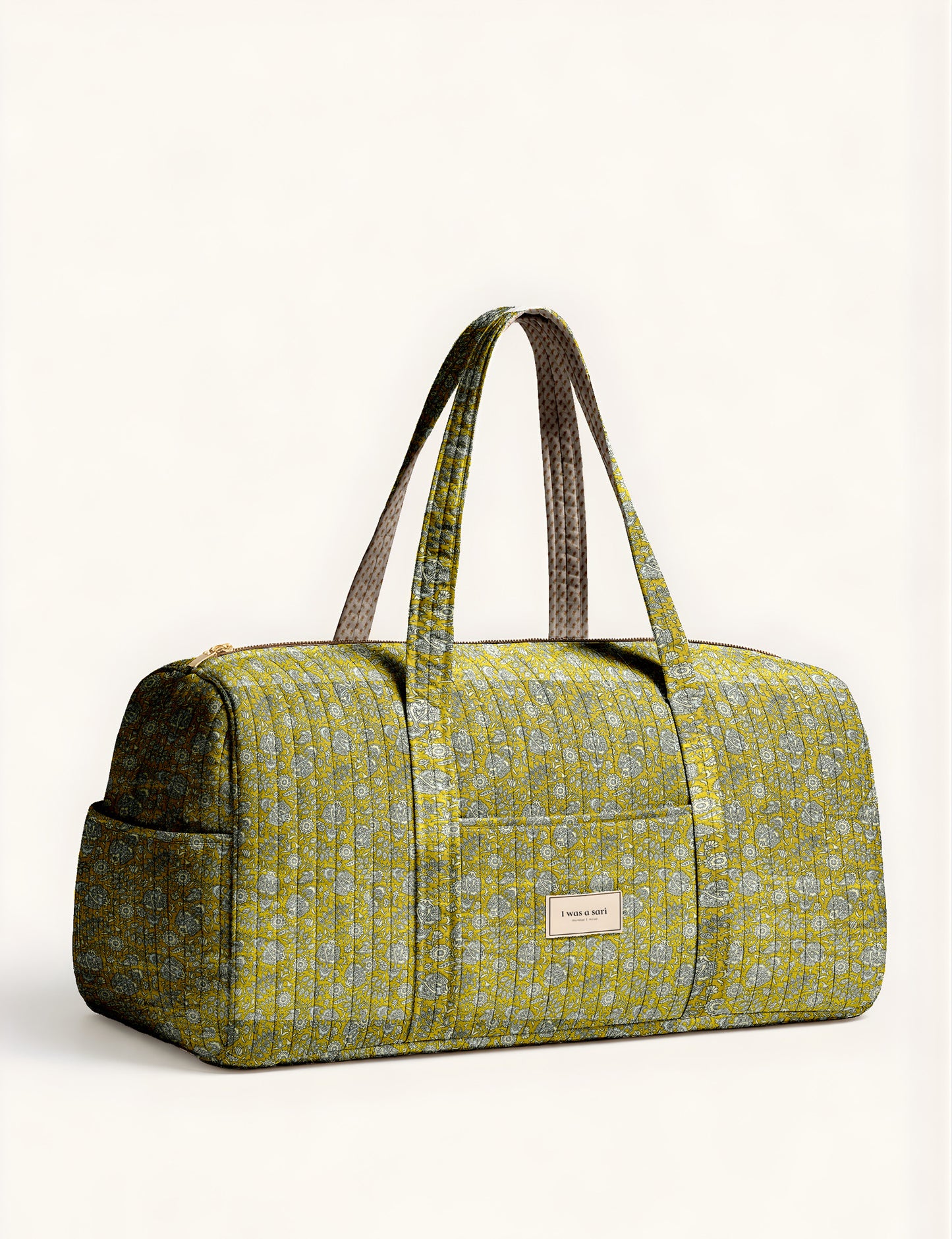Woman holding a brown paisley quilted barrel bag with front zipper pocket, wearing light blue jeans and white sneakers