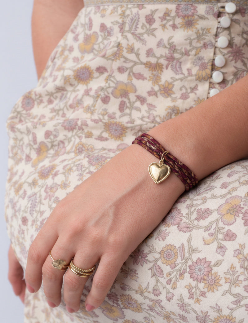 Red cord bracelet with gold heart charm worn on wrist, shown with floral patterned dress and gold rings on hand