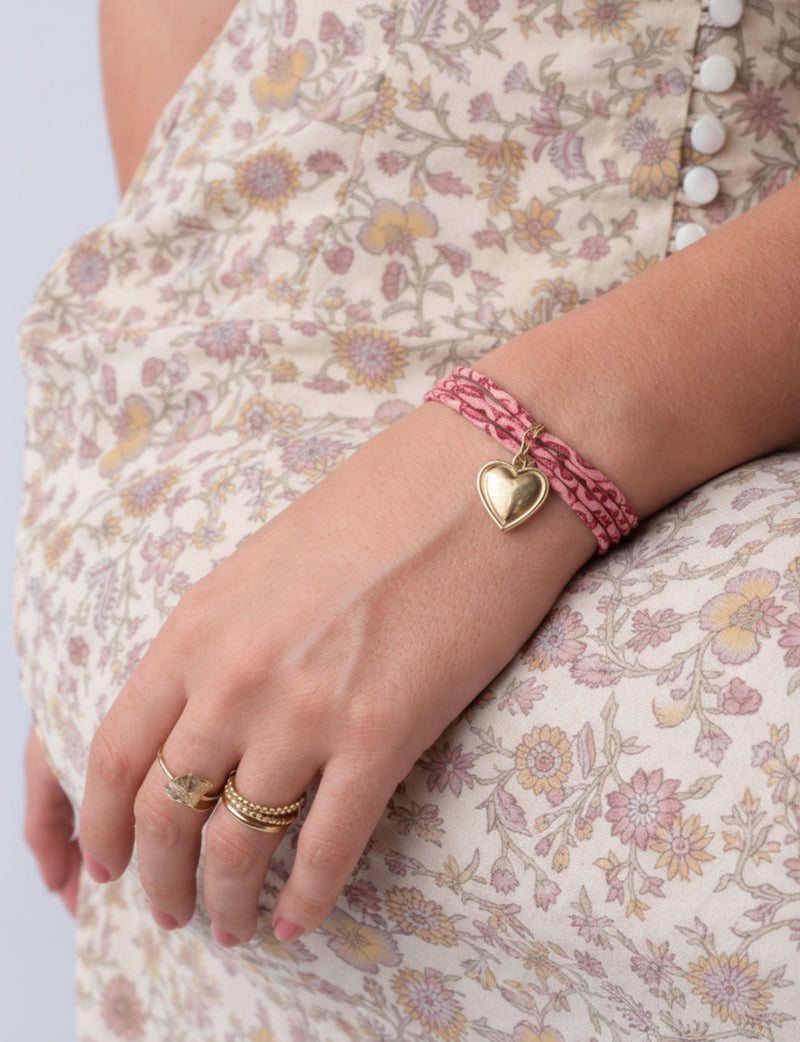 Pink cord bracelet with gold heart charm worn on wrist, shown with floral patterned dress and gold rings on hand