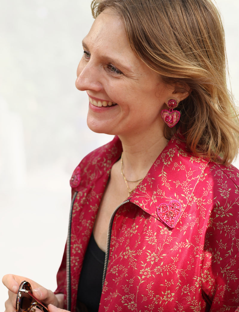 Woman wearing pink heart-shaped drop earrings with bead details, paired with a red floral patterned jacket and black top