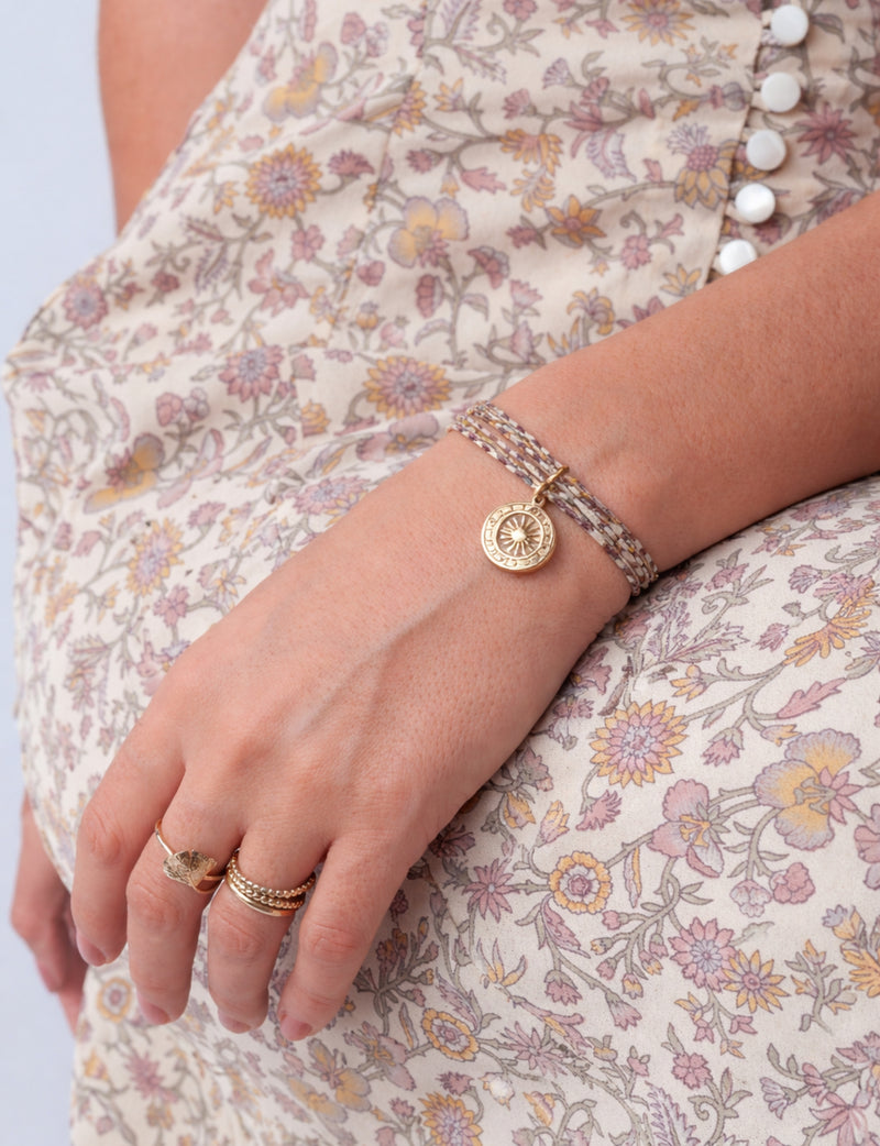 Beige and purple cord bracelet with gold sun medallion charm worn on wrist over floral patterned dress with white buttons