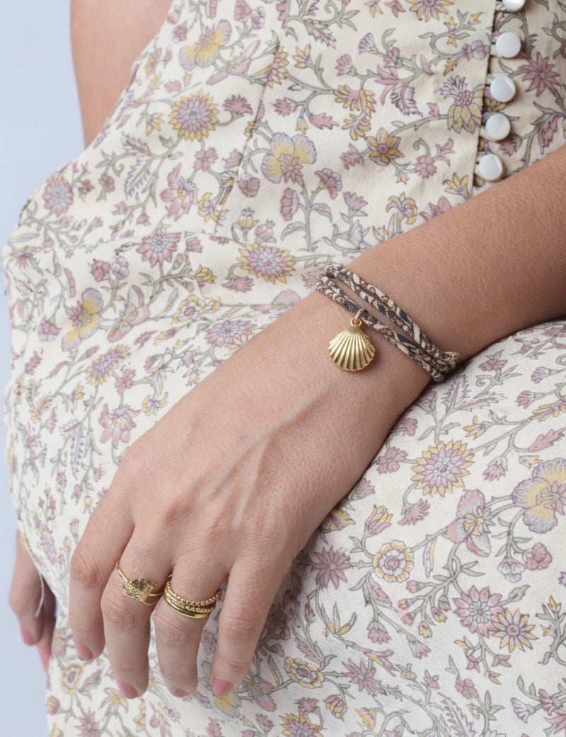 Beige cord bracelet with gold shell charm worn on wrist, paired with gold rings and floral patterned dress with white buttons