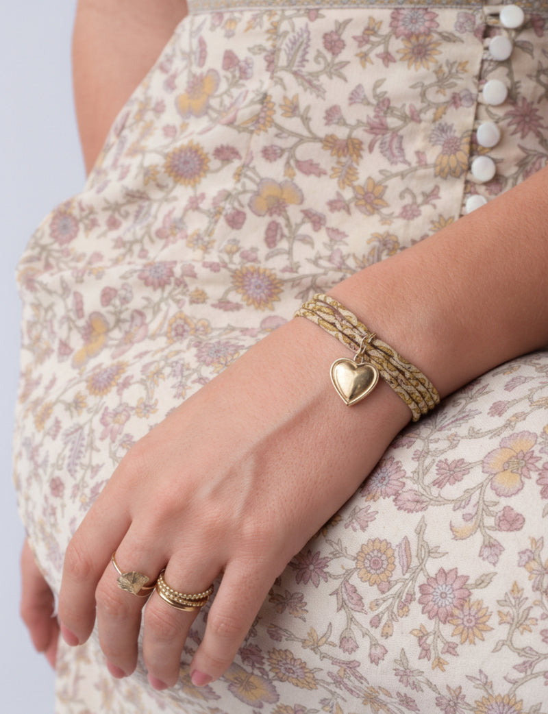 Close-up of a wrist wearing a gold cord bracelet with a heart charm, paired with gold rings on a floral dress