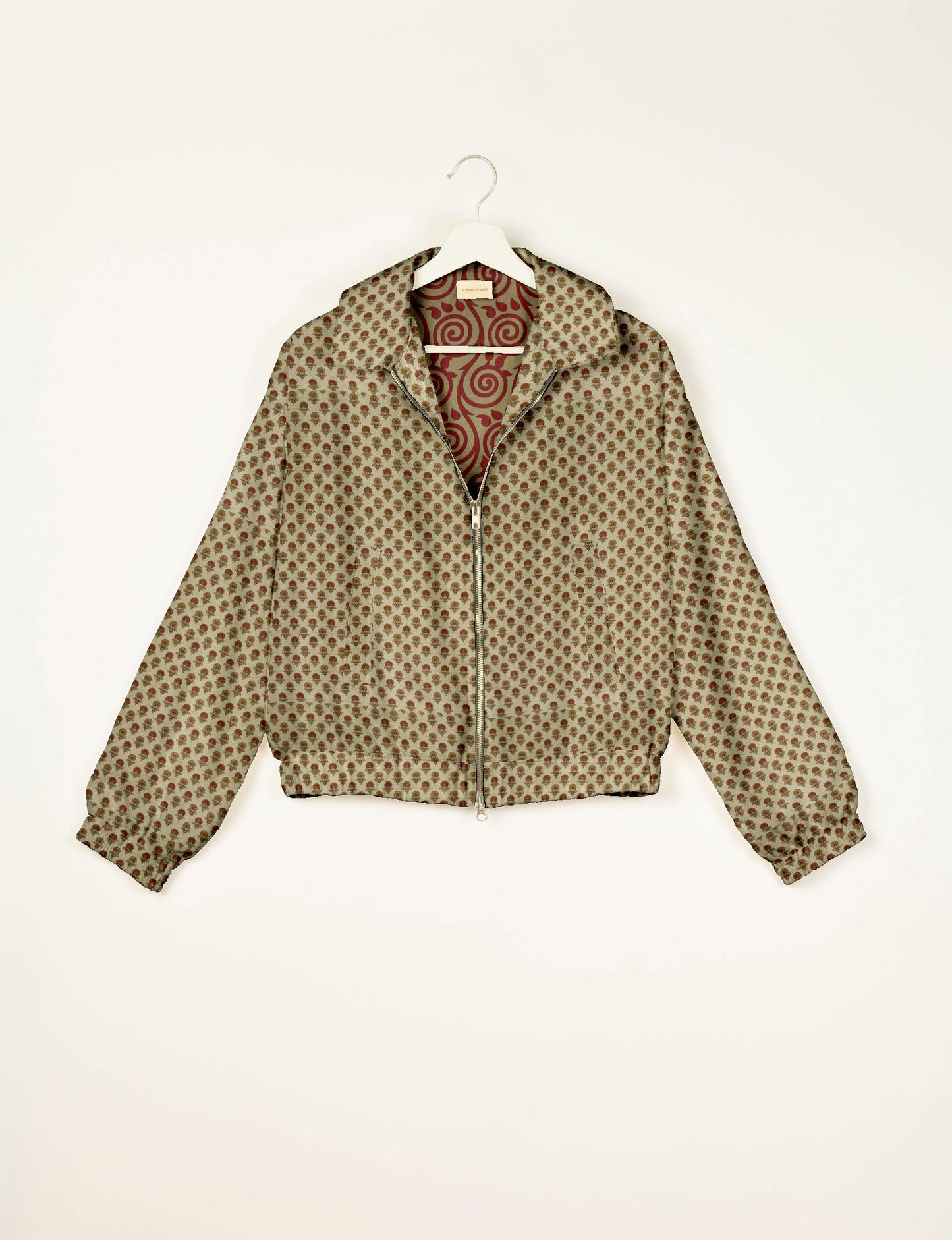 Beige bomber jacket with small brown floral pattern, front zipper, collar, elastic cuffs, and red swirl patterned lining, on