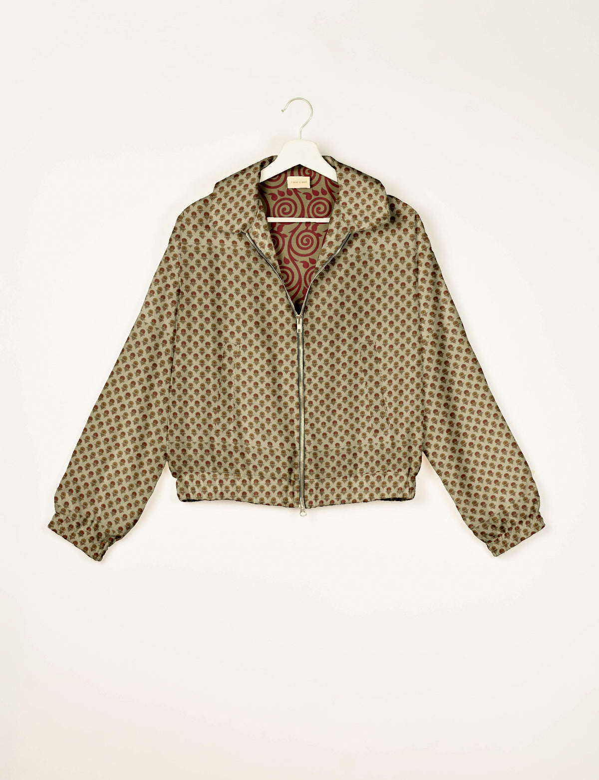 Beige bomber jacket with small brown floral pattern, front zipper, collar, elastic cuffs, and red swirl patterned lining, on