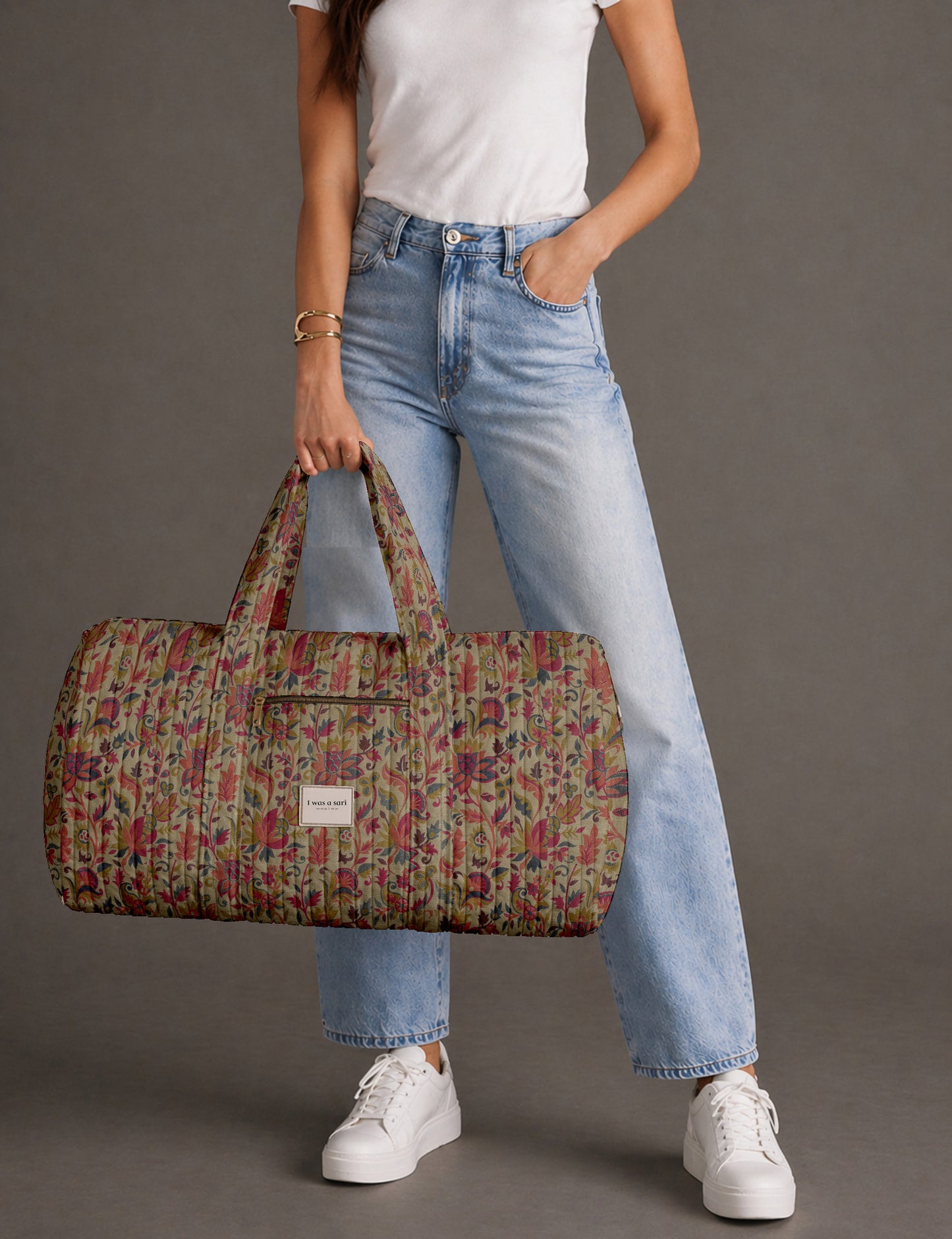 Woman holding a brown paisley quilted barrel bag with front zipper pocket, wearing light blue jeans and white sneakers