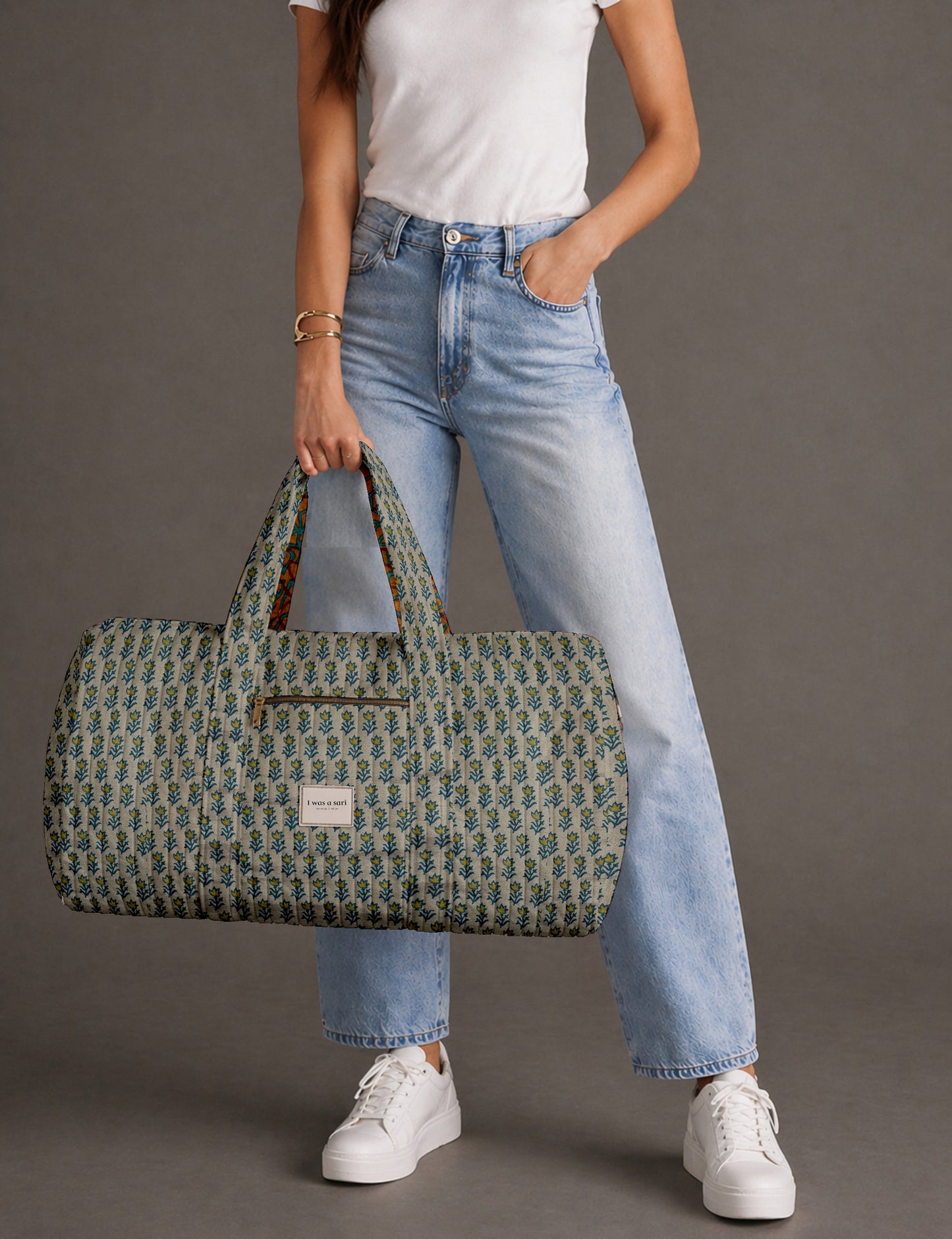 Woman holding a brown paisley quilted barrel bag with front zipper pocket, wearing light blue jeans and white sneakers
