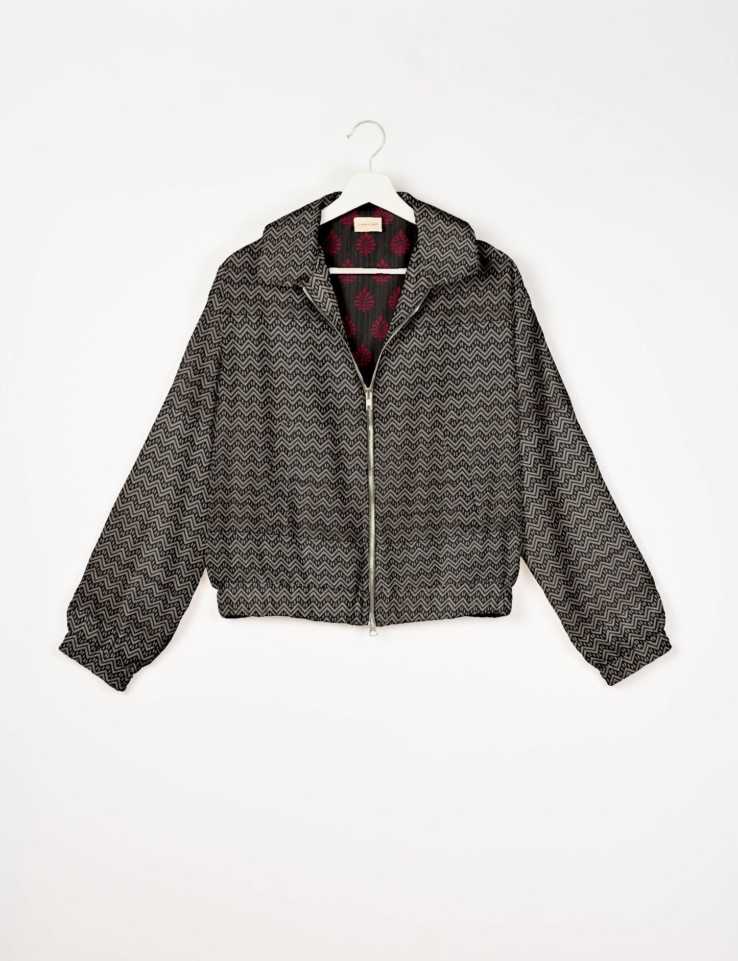 Beige bomber jacket with black geometric zigzag pattern, front zipper, elastic cuffs and hem, shown on hanger