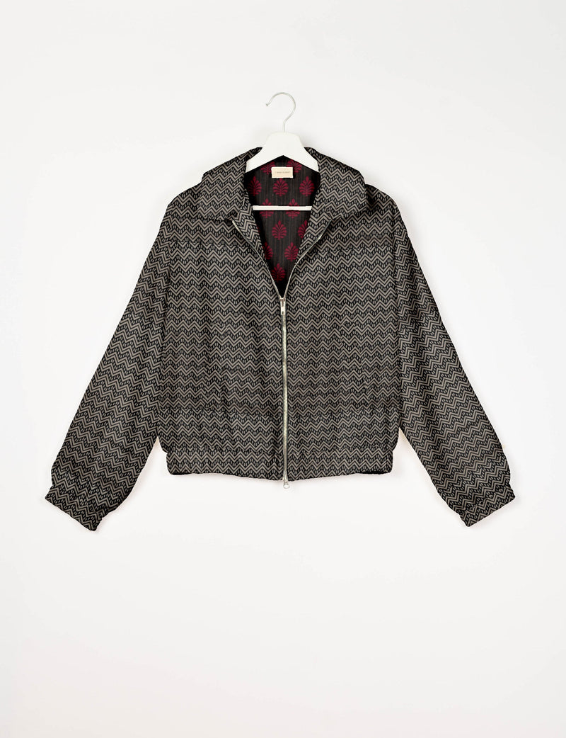 Beige bomber jacket with black geometric zigzag pattern, front zipper, elastic cuffs and hem, shown on hanger