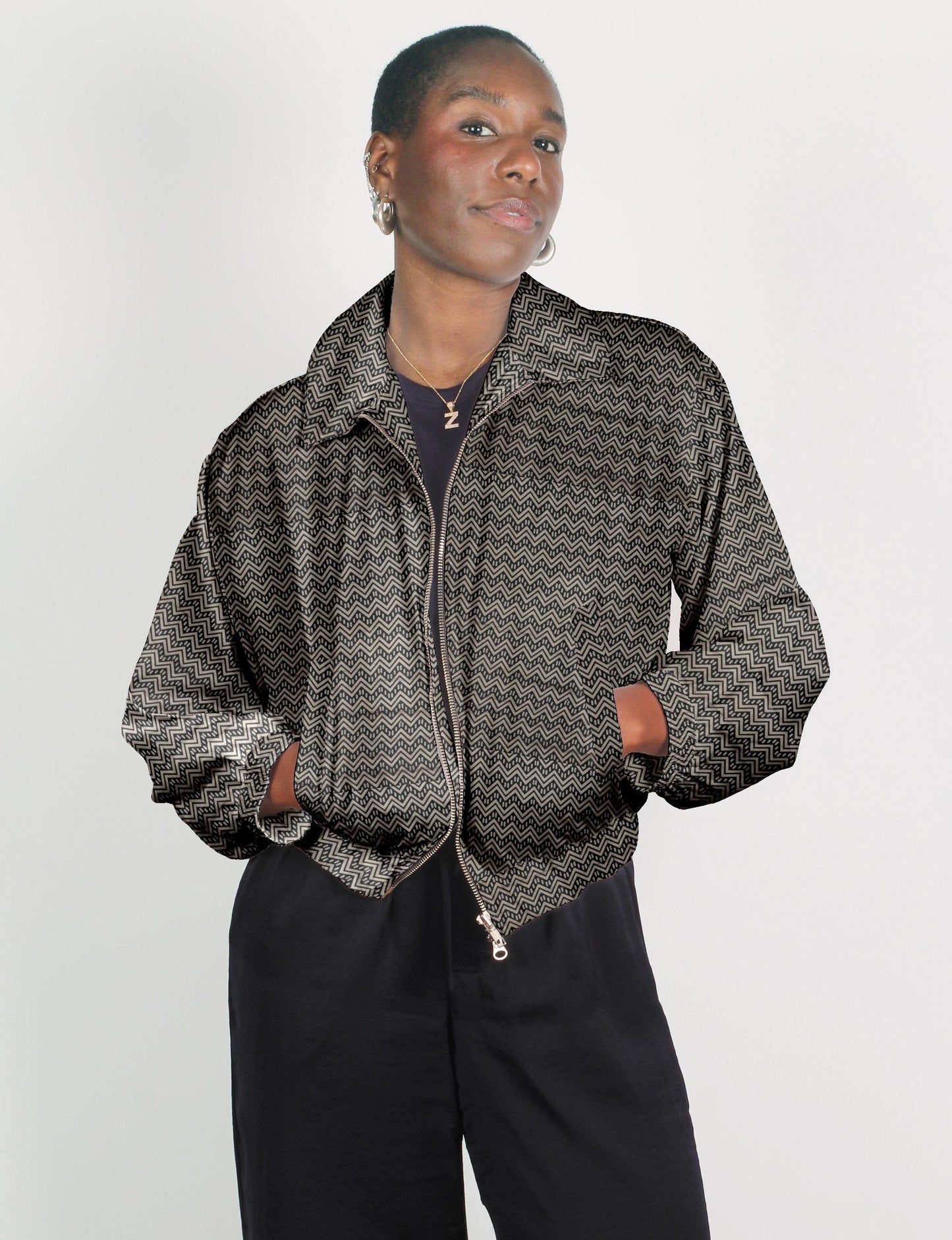 Woman wearing beige bomber jacket with black geometric zigzag pattern, front zipper, and pockets, paired with black pants