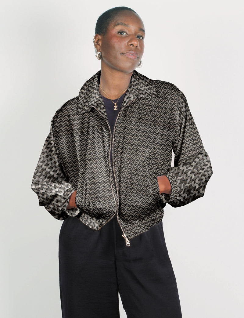 Woman wearing beige bomber jacket with black geometric zigzag pattern, front zipper, and pockets, paired with black pants
