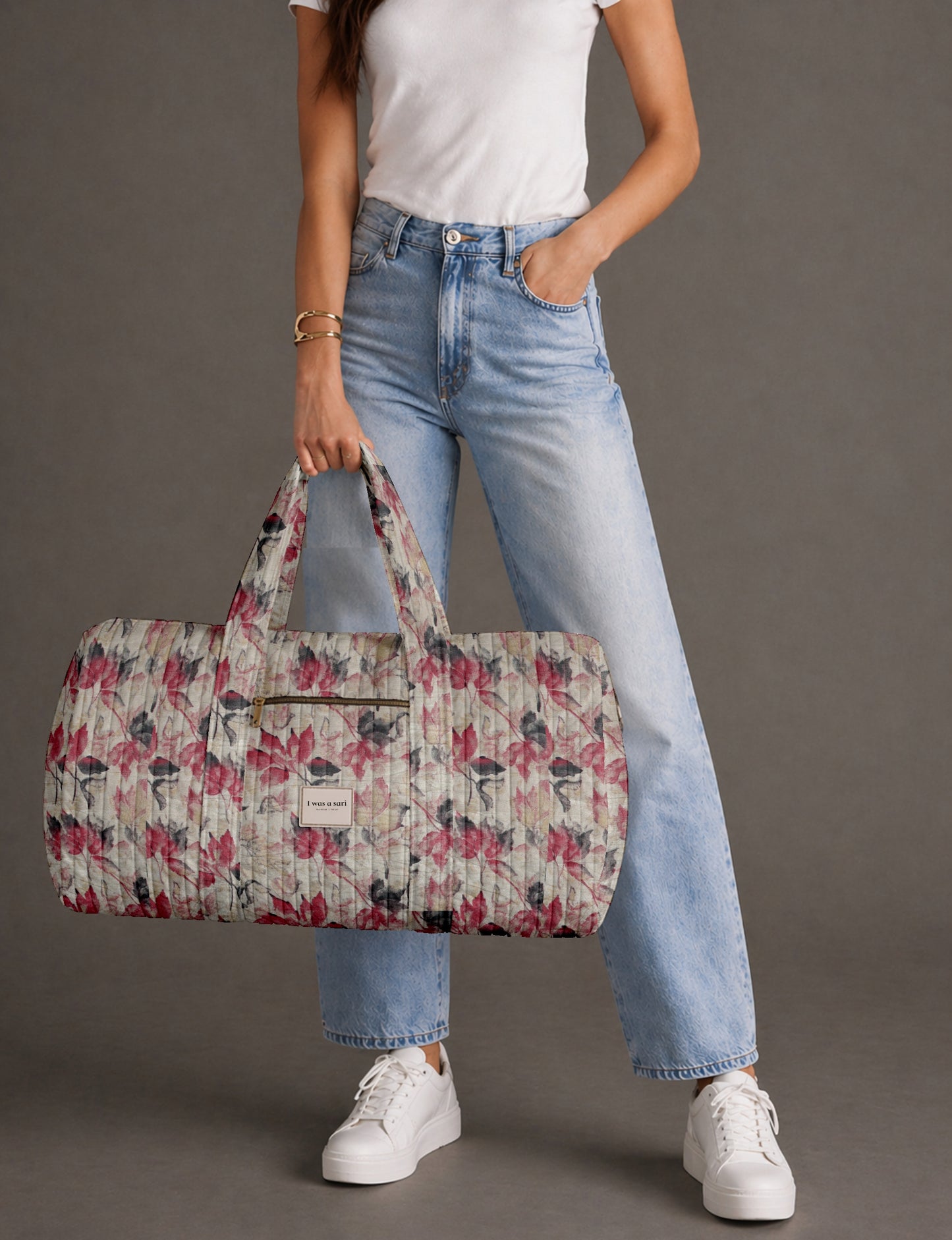 Woman wearing white t-shirt and light blue jeans holding a beige quilted barrel bag with red and black leaf pattern and
