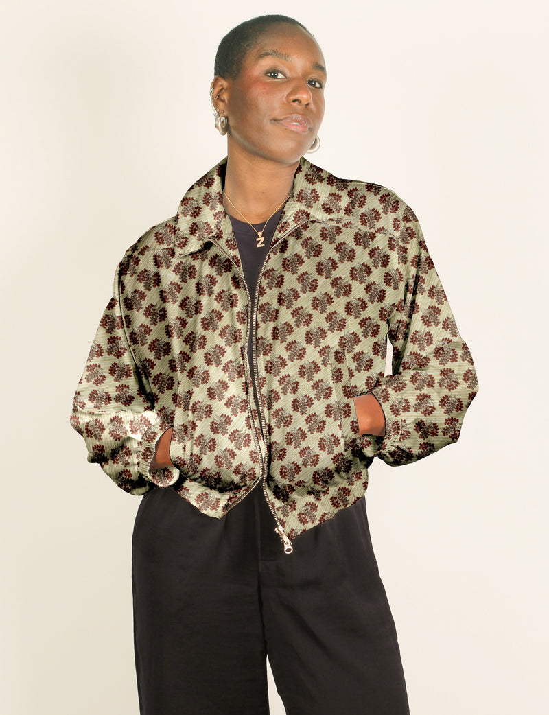 Woman wearing beige bomber jacket with brown leaf pattern, front zipper, and elastic cuffs, paired with black pants
