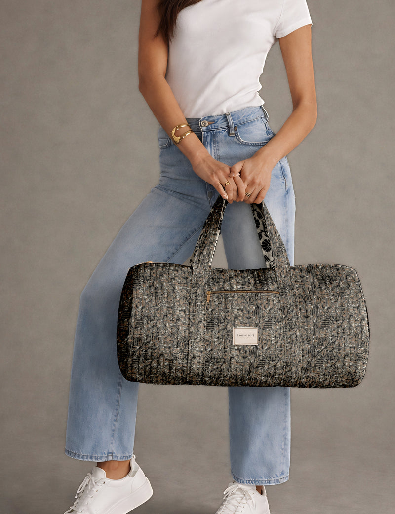 Quilted black abstract bowling bag with front zipper pocket, held by person wearing white top and light blue jeans