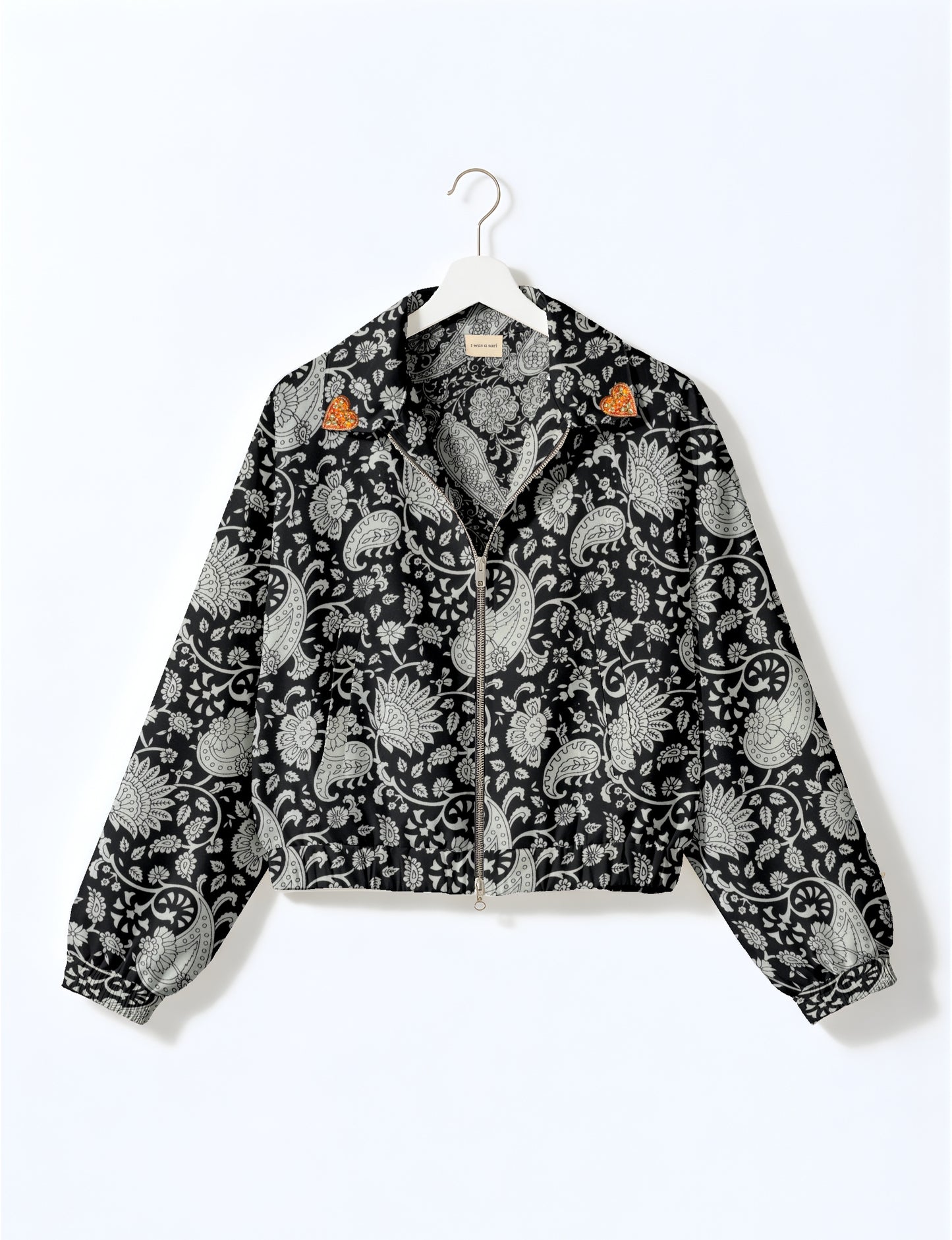Black paisley bomber jacket with front zipper, elastic cuffs and hem, and gold heart patches on collar tips hanging on white