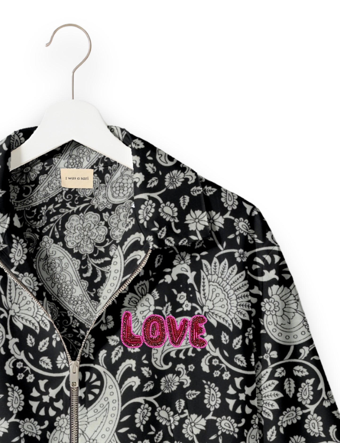 Black bomber jacket with white paisley floral pattern, front zipper, and pink beaded "LOVE" patch on chest, shown on white