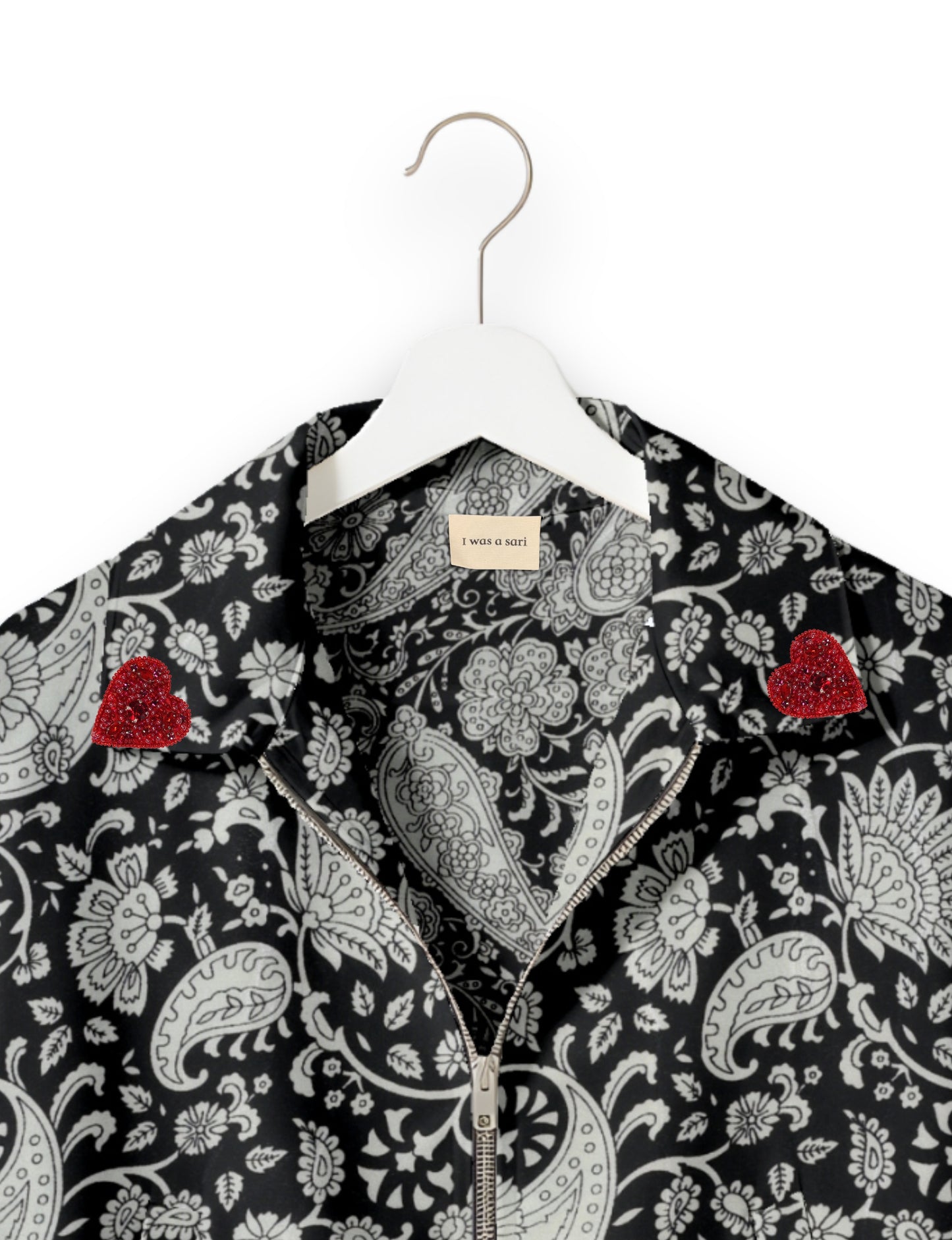 Black bomber jacket with white paisley floral pattern, silver zipper, red beaded heart patches on collar, on white hanger