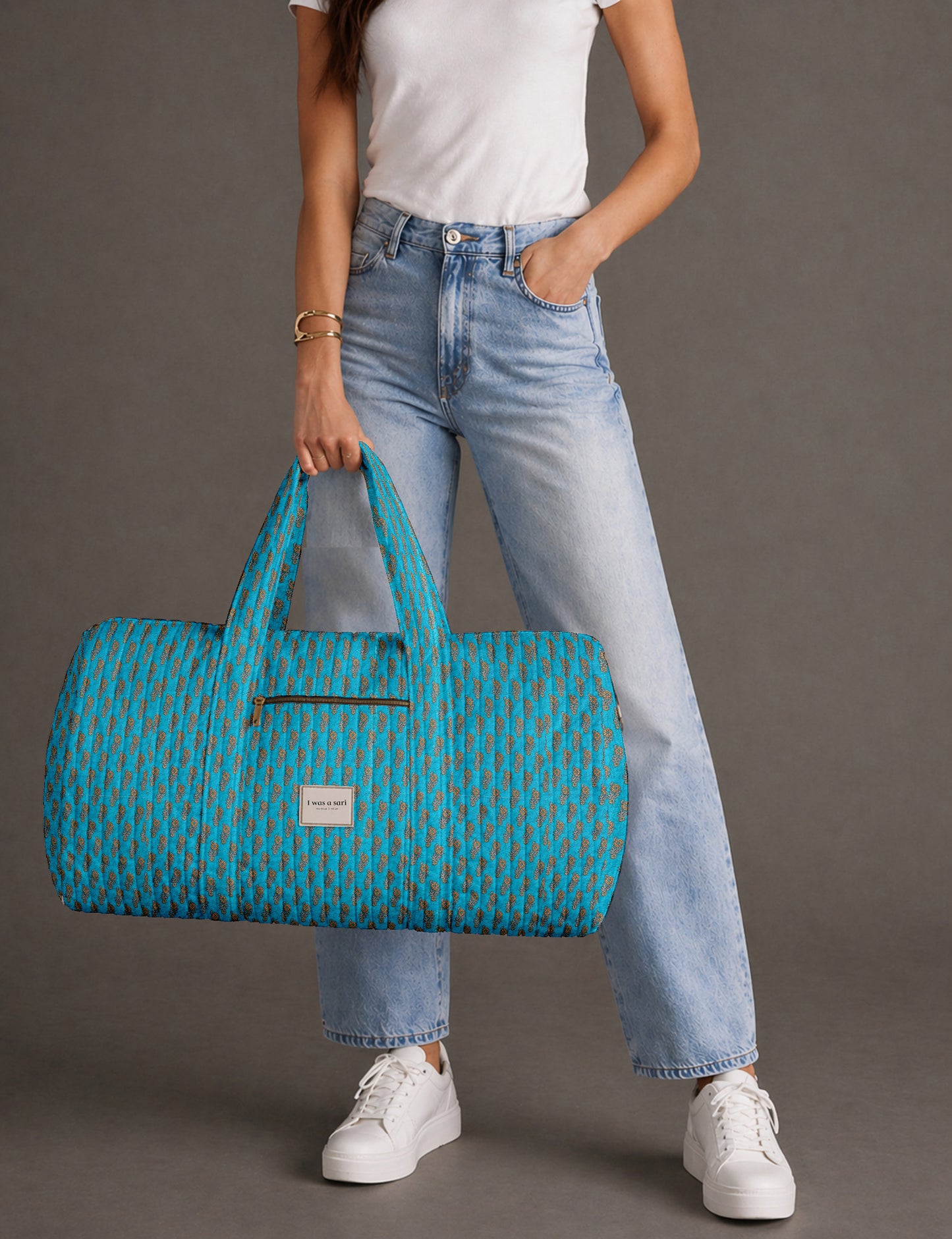 Woman holding a brown paisley quilted barrel bag with front zipper pocket, wearing light blue jeans and white sneakers