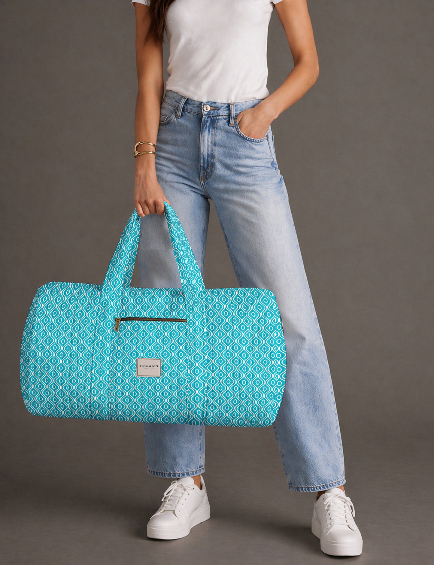 Woman holding a brown paisley quilted barrel bag with front zipper pocket, wearing light blue jeans and white sneakers