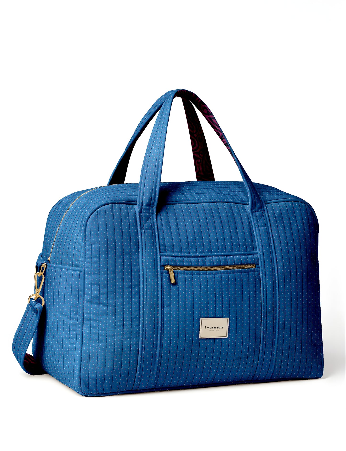 Blue quilted duffle bag with micro dot pattern, front zip pocket, dual handles, and "I was a sari" label, side strap visible