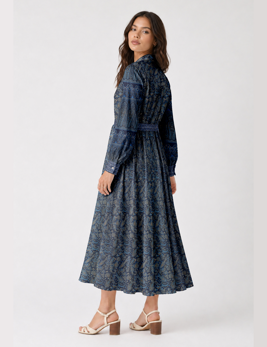 Woman wearing a long blue paisley shirt dress with long sleeves and buttoned cuffs, shown from back side angle