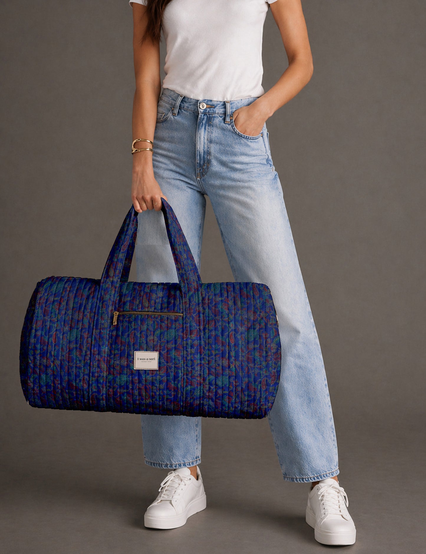 Woman holding a brown paisley quilted barrel bag with front zipper pocket, wearing light blue jeans and white sneakers