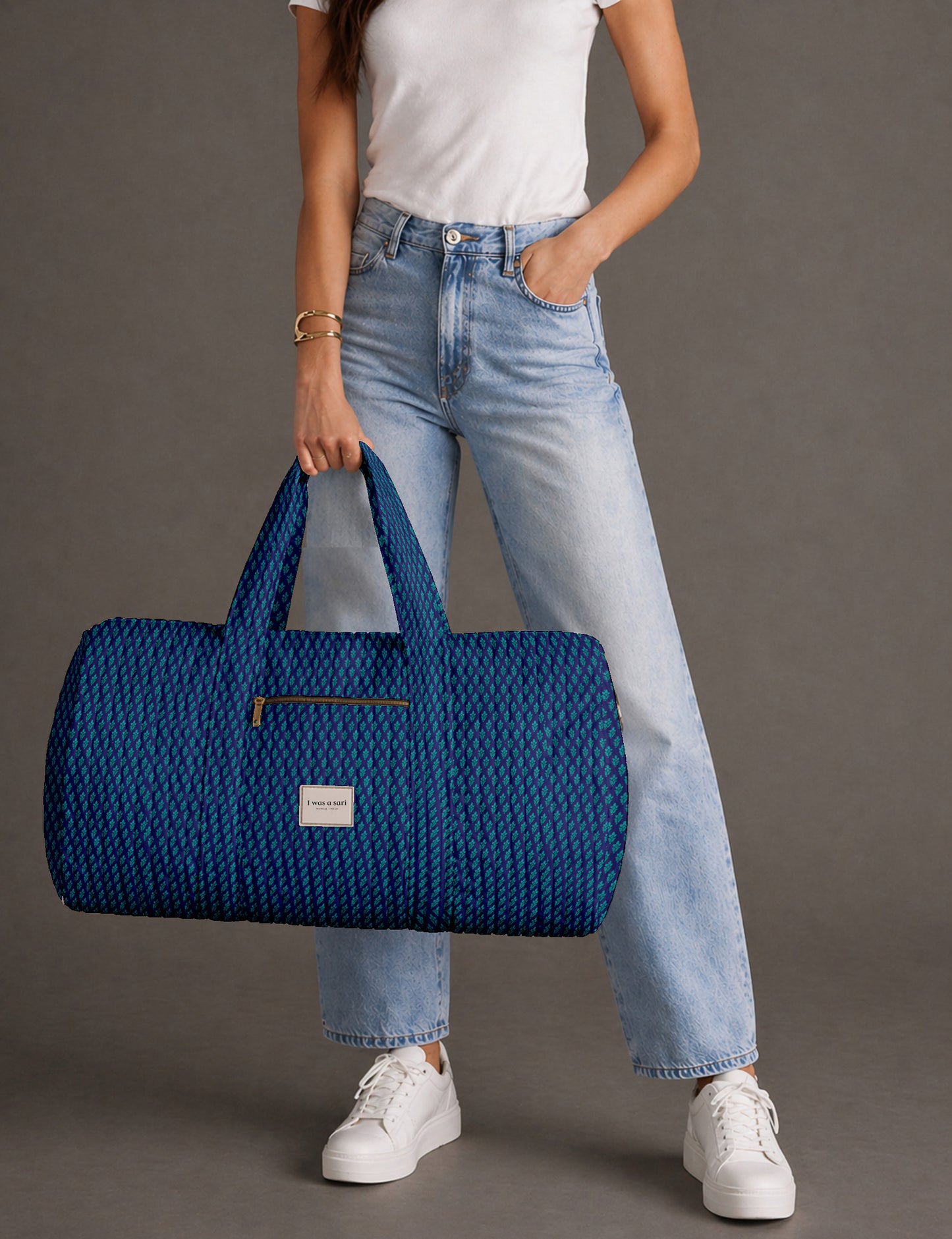 Woman holding a brown paisley quilted barrel bag with front zipper pocket, wearing light blue jeans and white sneakers