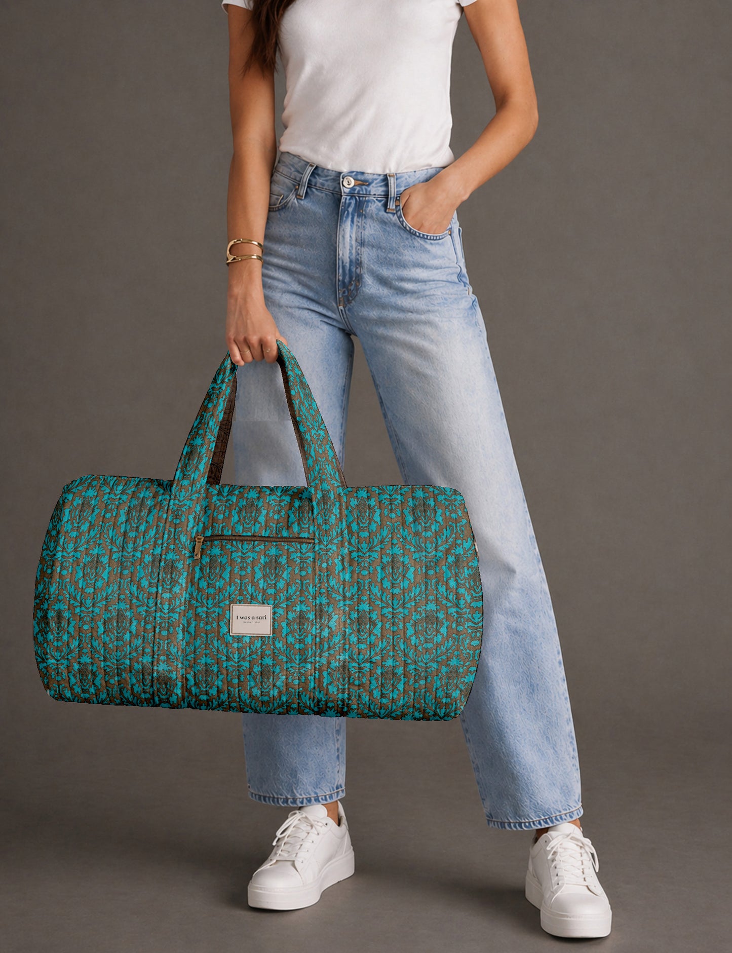 Woman holding a brown paisley quilted barrel bag with front zipper pocket, wearing light blue jeans and white sneakers