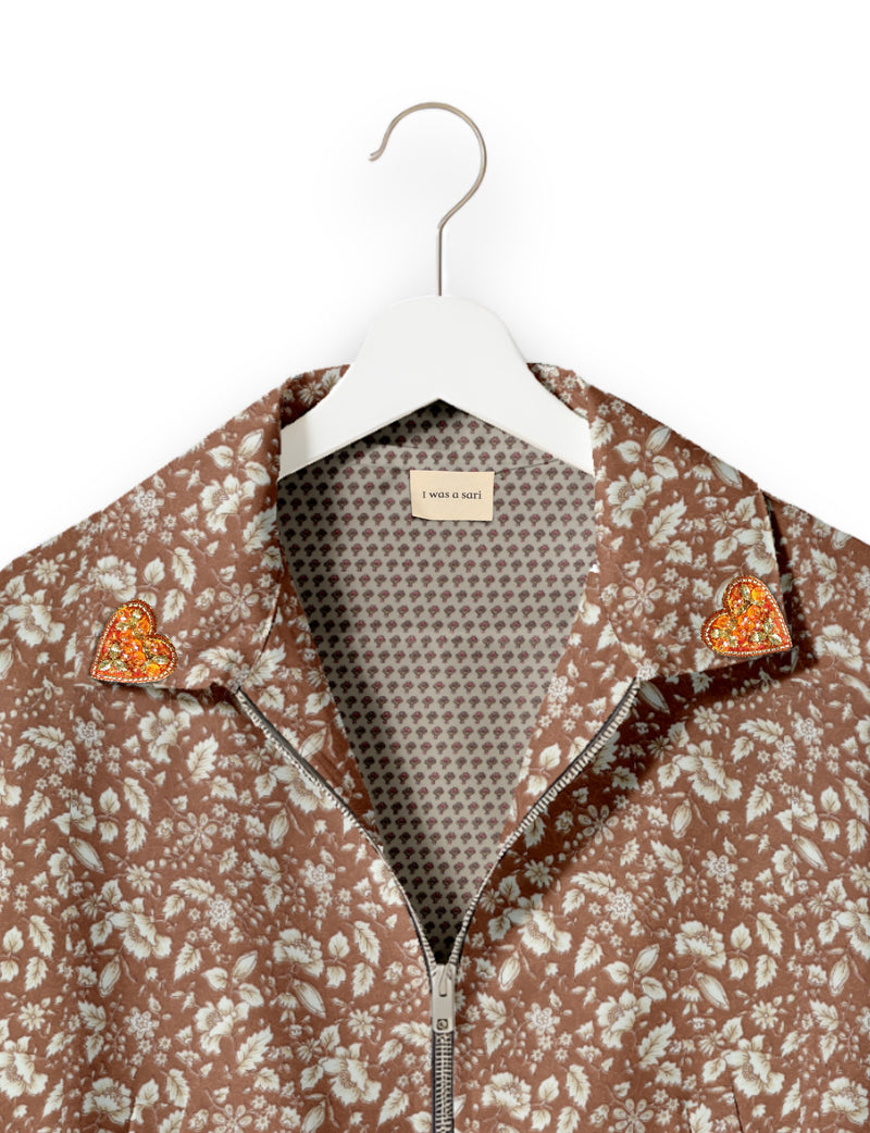 Brown floral bomber jacket with silver zipper and orange jeweled heart pins on collar, shown on white hanger