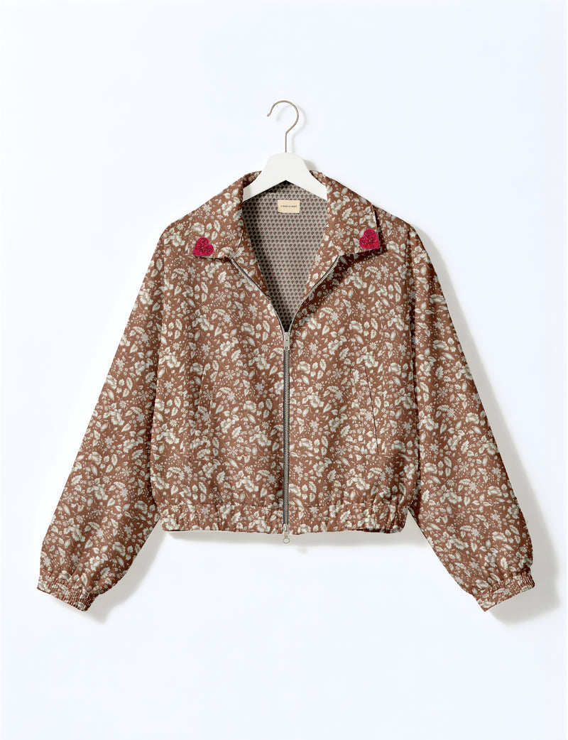 Brown floral bomber jacket with front zipper, elastic cuffs and hem, and pink heart patches on collar tips hanging on white hanger