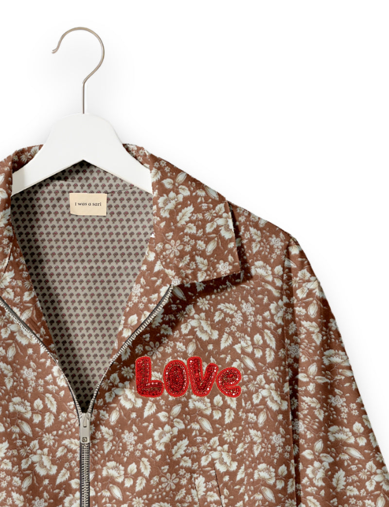Brown floral bomber jacket with red beaded "LOVE" patch, silver zipper, and patterned inner lining on white hanger