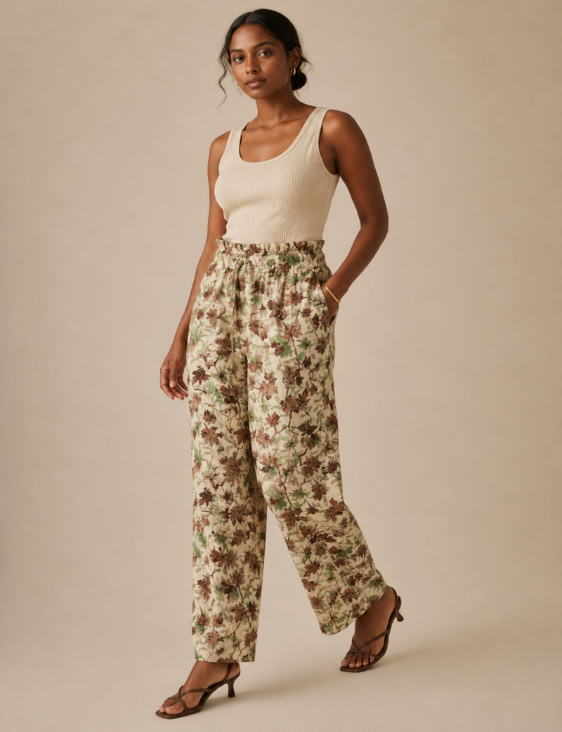 A woman modeling a pair of high-waisted, wide-leg trousers with a brown and green floral pattern on a cream background, paired with a cream tank top and brown strappy heels, against a solid beige studio background.