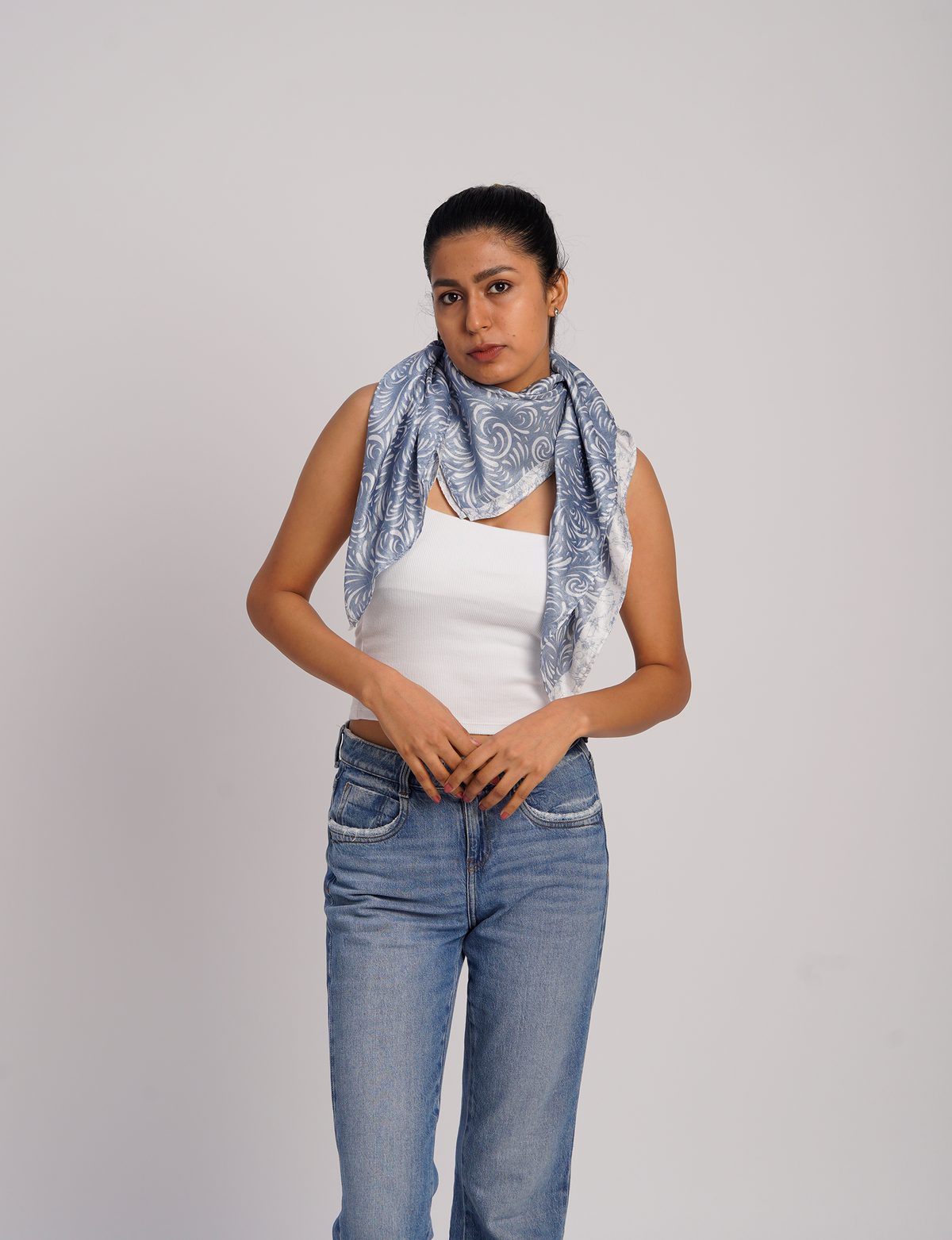 Woman wearing a blue and white patterned foulard scarf draped around her neck, paired with a white tank top and blue jeans