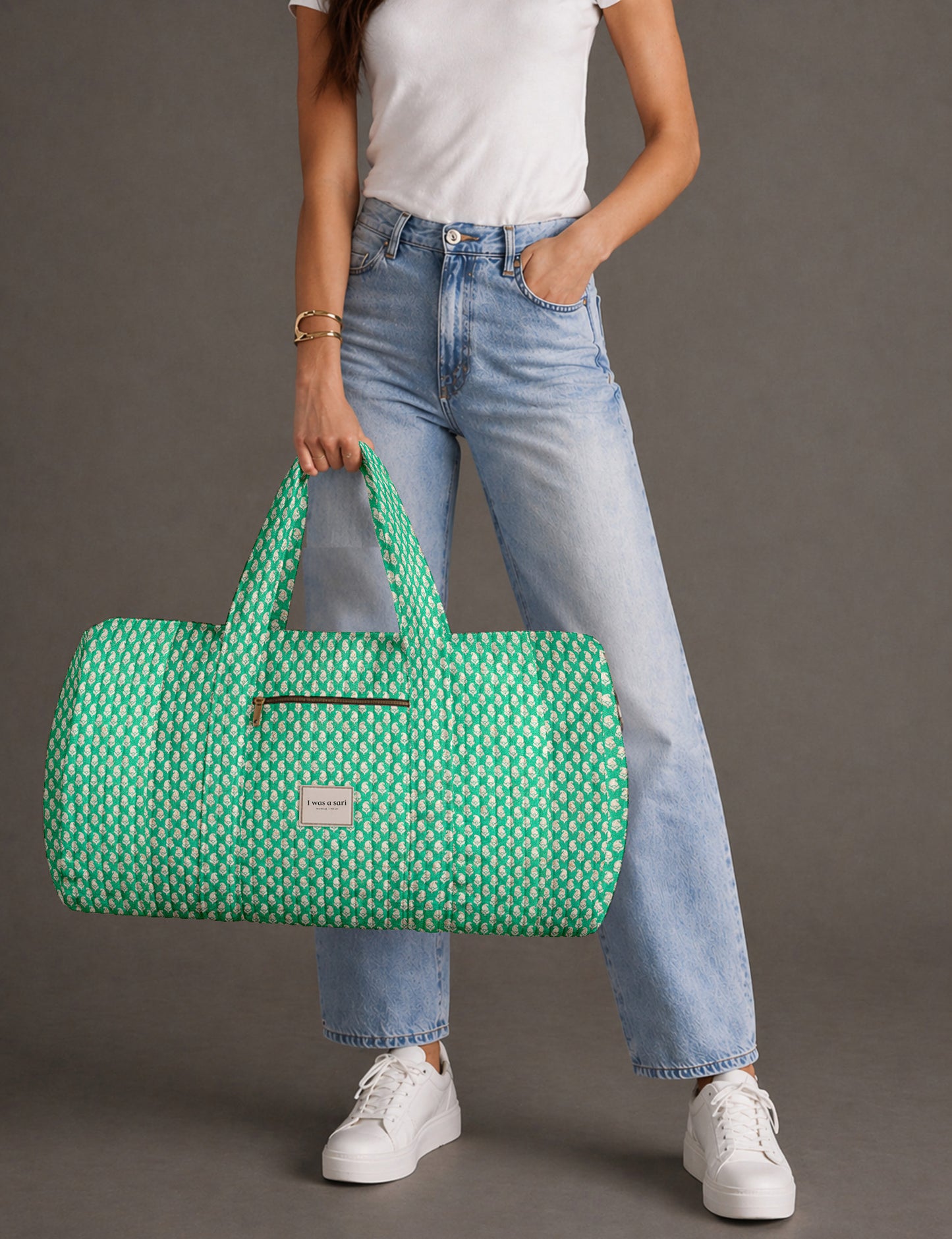 Woman holding a brown paisley quilted barrel bag with front zipper pocket, wearing light blue jeans and white sneakers