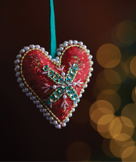 Red heart-shaped ornament with beaded snowflake design and pearl trim hanging by green ribbon against dark background