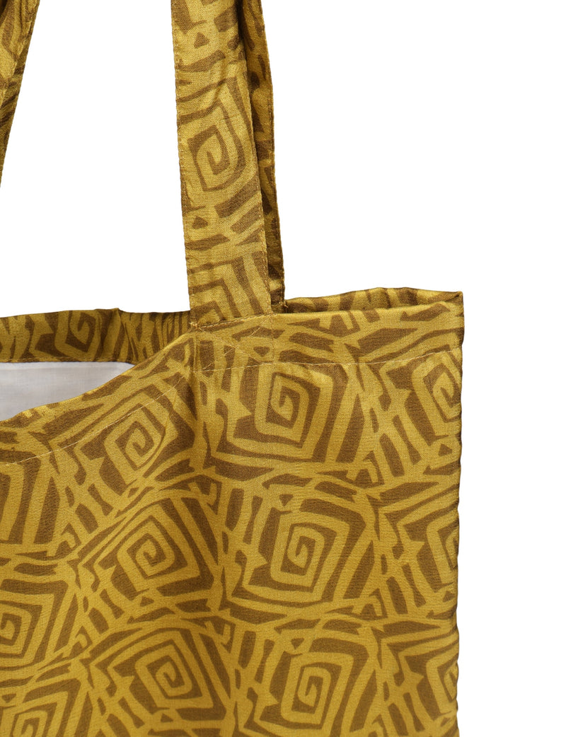 Yellow shopper bag with dark yellow geometric spiral pattern, fabric texture visible, showing top corner and strap close-up