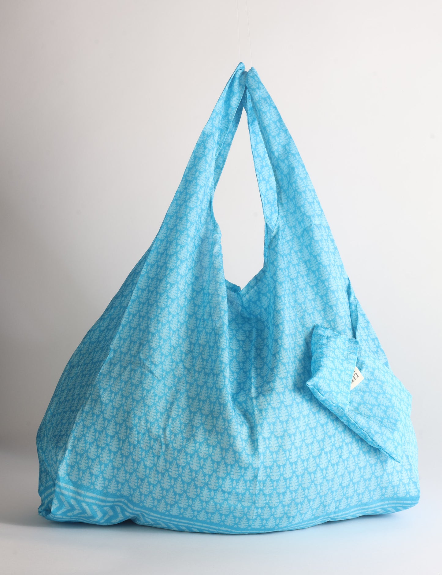 Blue reusable bag with tropical floral pattern in pink, yellow, and green, shown hanging with handles visible