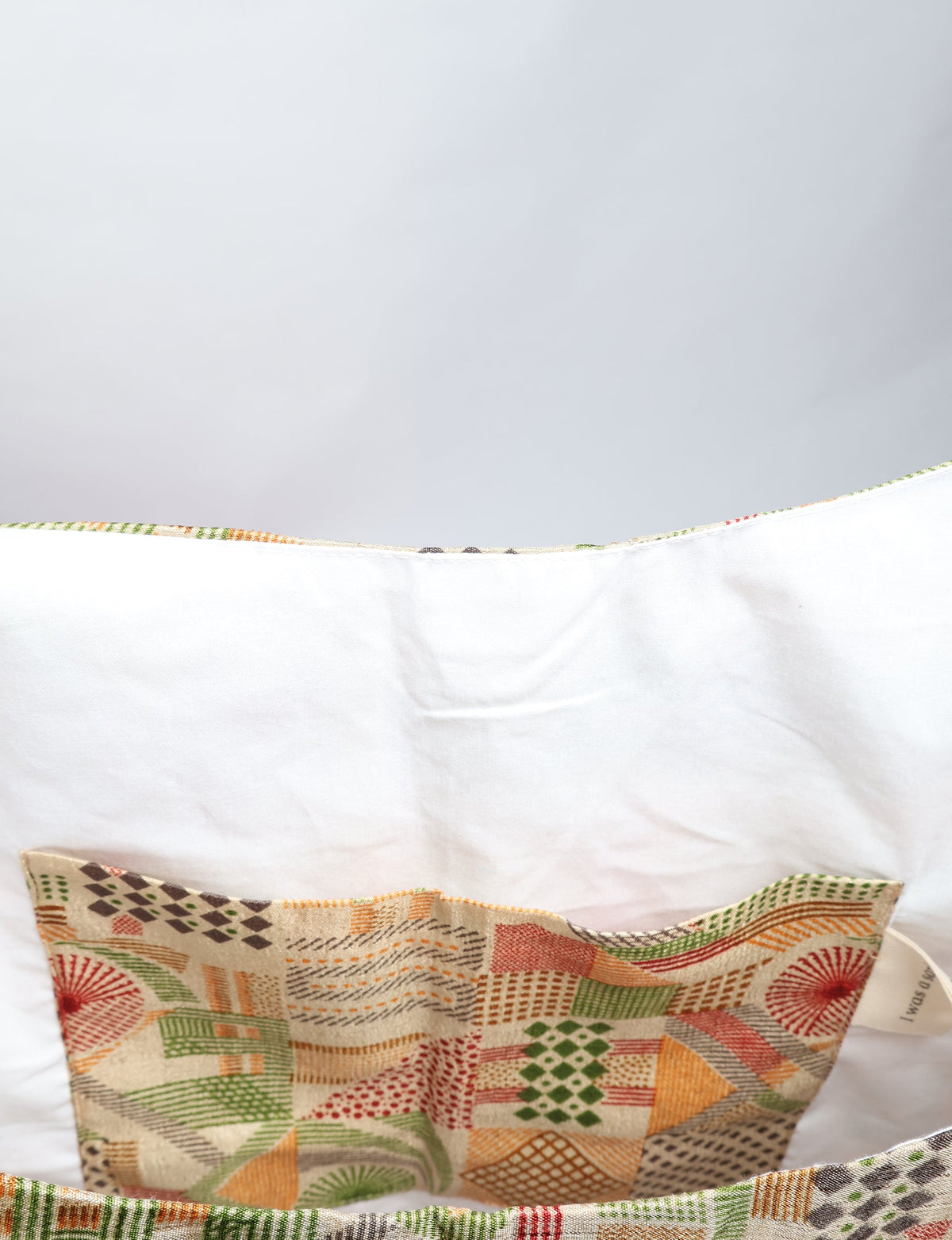 Beige trapeze bag interior with white lining and a geometric patterned fabric pocket in green, orange, and red tones