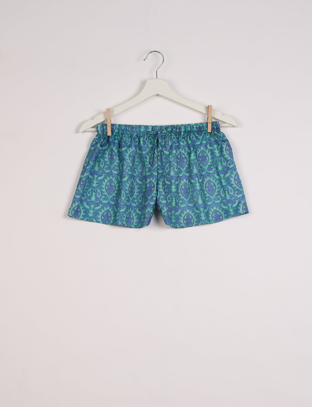 Blue and green Indian ethnic patterned shorts with elastic waistband hanging on white hanger against white background