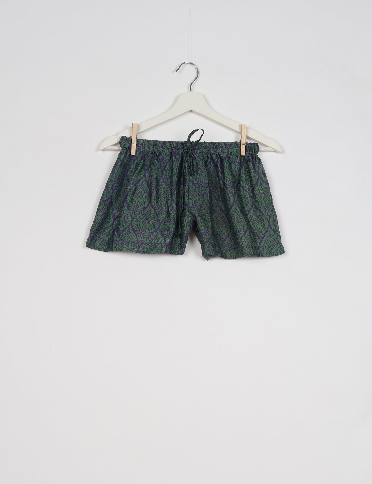 Purple shorts with green leaf pattern, elastic waistband with drawstring, hanging on white hanger against white background