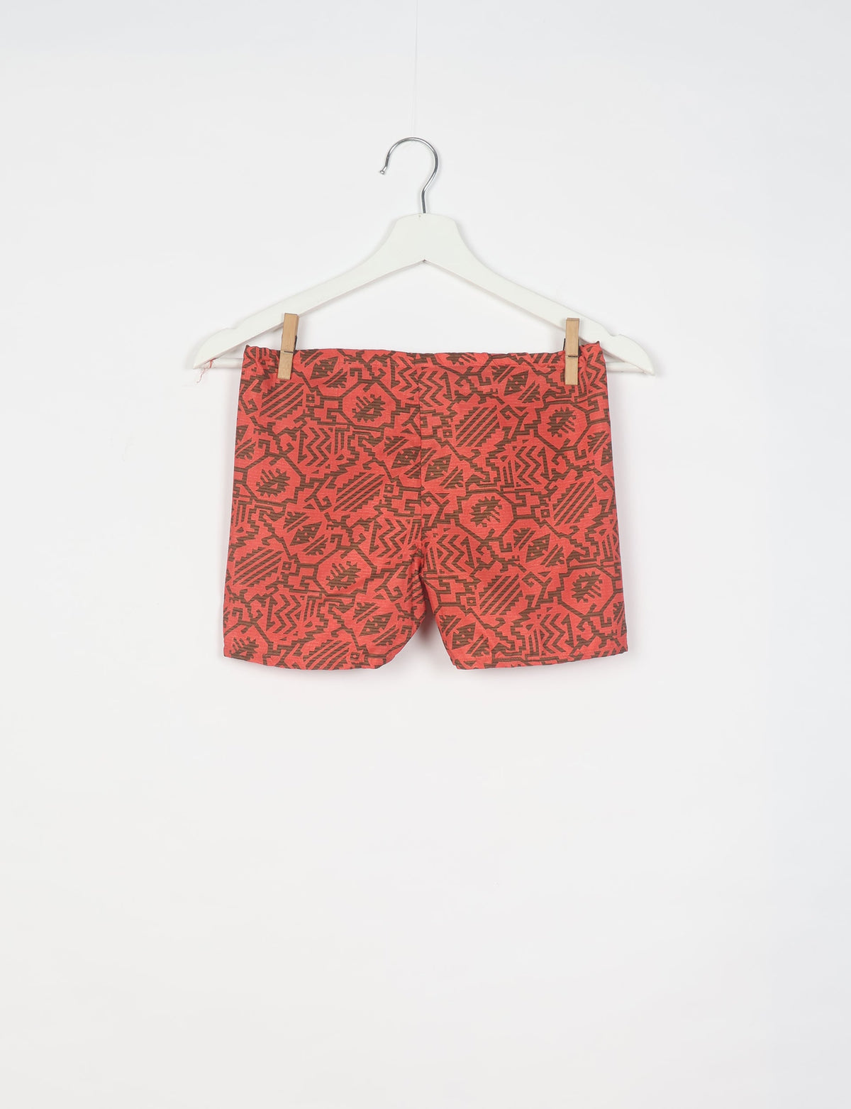 Orange shorts with dark brown tropical geometric pattern hanging on white hanger against white background