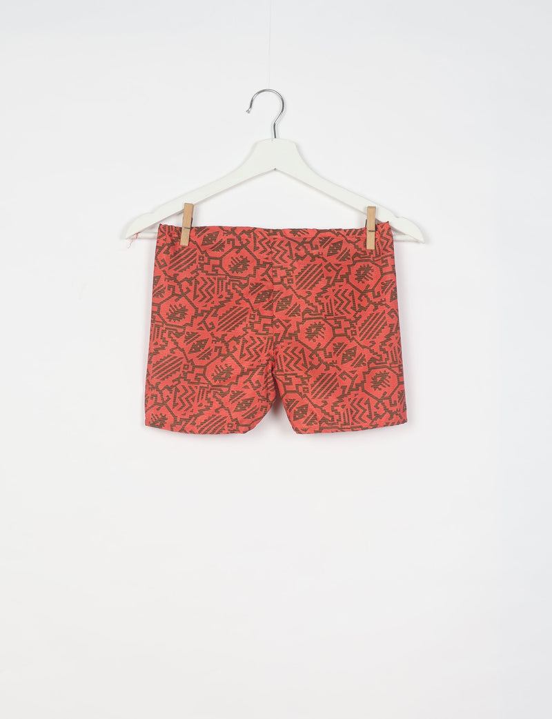 Orange shorts with dark brown tropical geometric pattern hanging on white hanger against white background