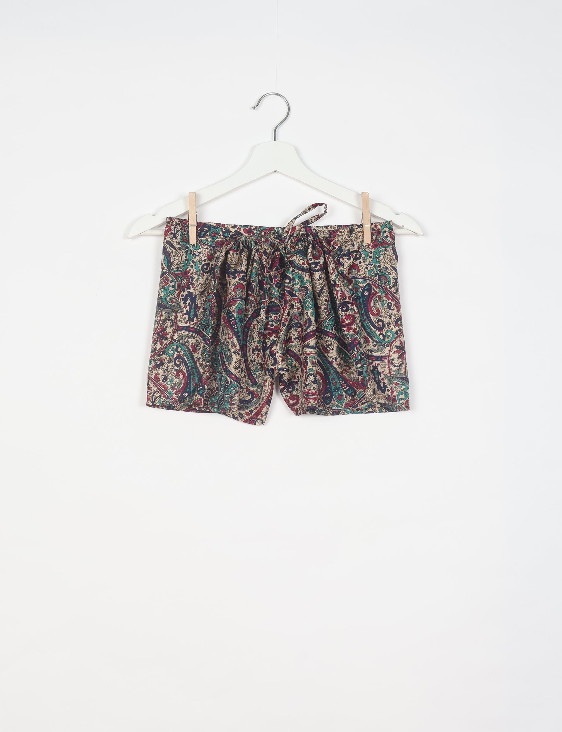 Beige tropical print shorts with red, green, and navy paisley pattern, elastic waistband, and drawstring, hanging on white