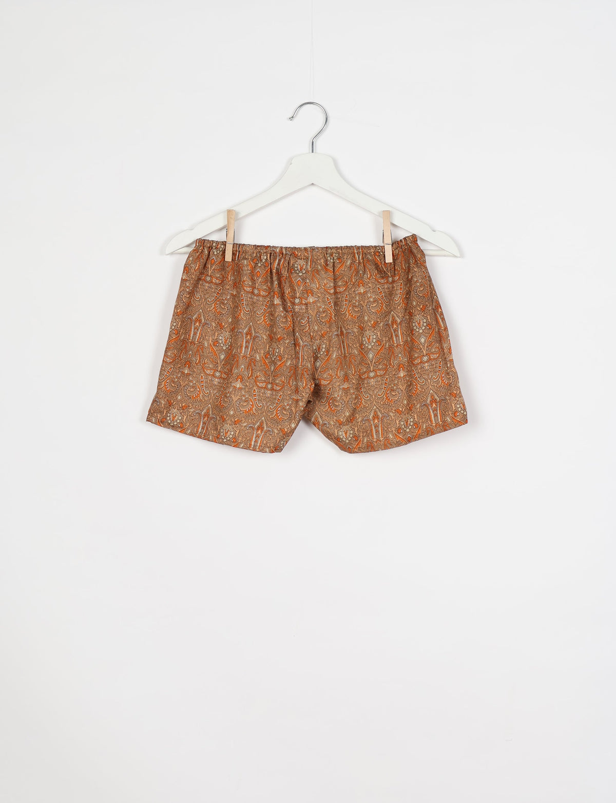 Brown and orange paisley patterned shorts with elastic waistband hanging on white hanger against white background