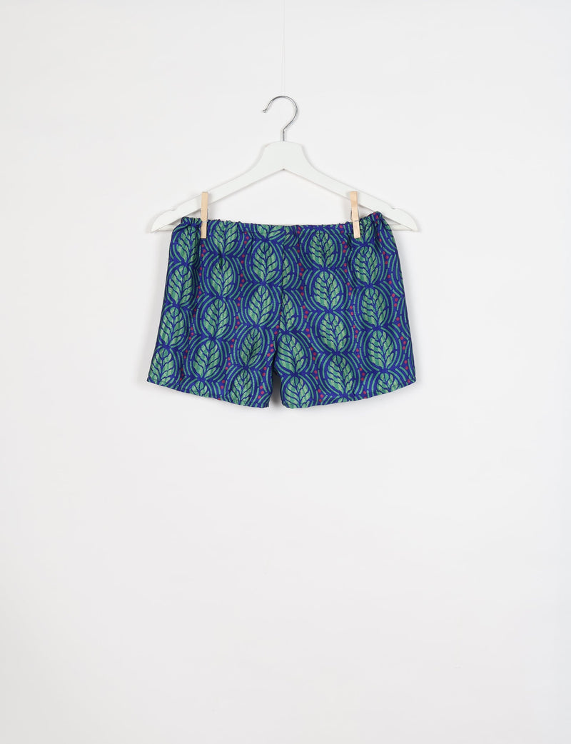 Blue shorts with green leaf pattern and small red dots, elastic waistband, hanging on white hanger against white background